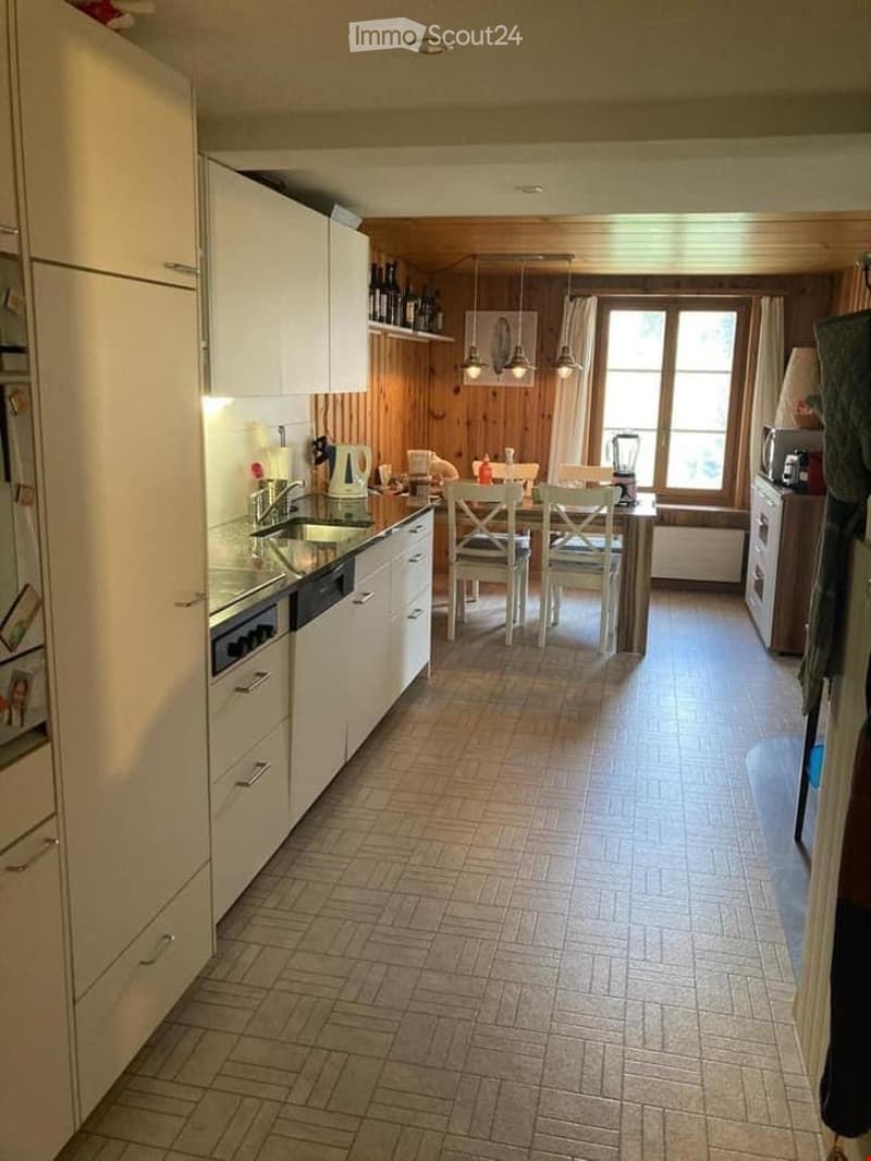 Modern kitchen with white cabinets, a central island, and a dining area. Features include a refrigerator, sink, oven, dishwasher, blender, and a table with chairs. Wooden walls and a tiled floor complete the space.