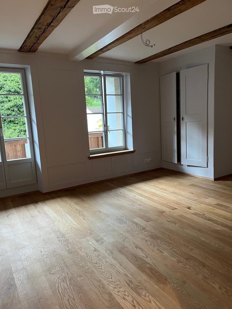 Empty room with wooden flooring, large windows, and exposed wooden beams on the ceiling.