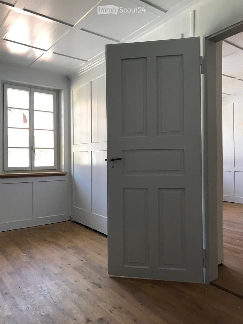Empty room with light grey walls, wooden floor, and a grey door leading to another room. A window with multiple panes is visible on the left side.
