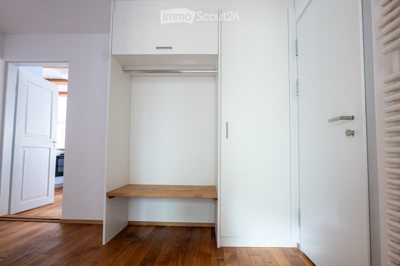 Empty room with white walls, wooden floor, built-in closet with wooden shelf, and a door leading to another room.
