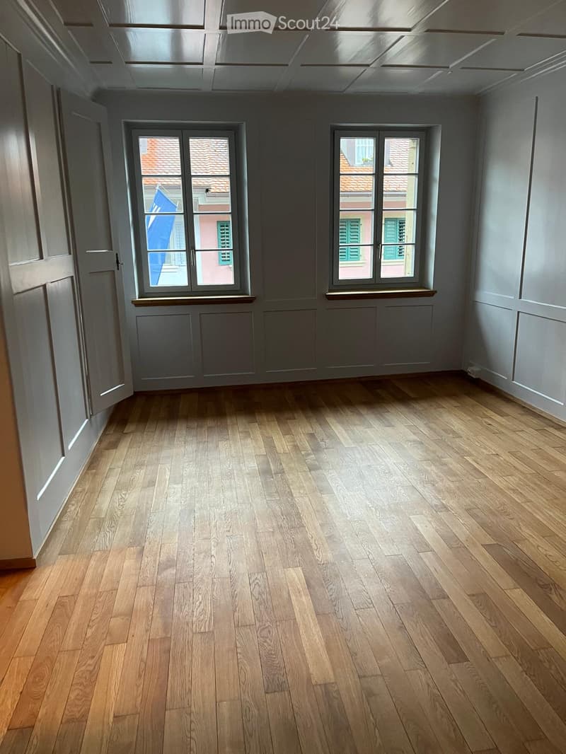 Empty room with wooden flooring, grey walls, and two windows overlooking a tiled roof and neighboring building.