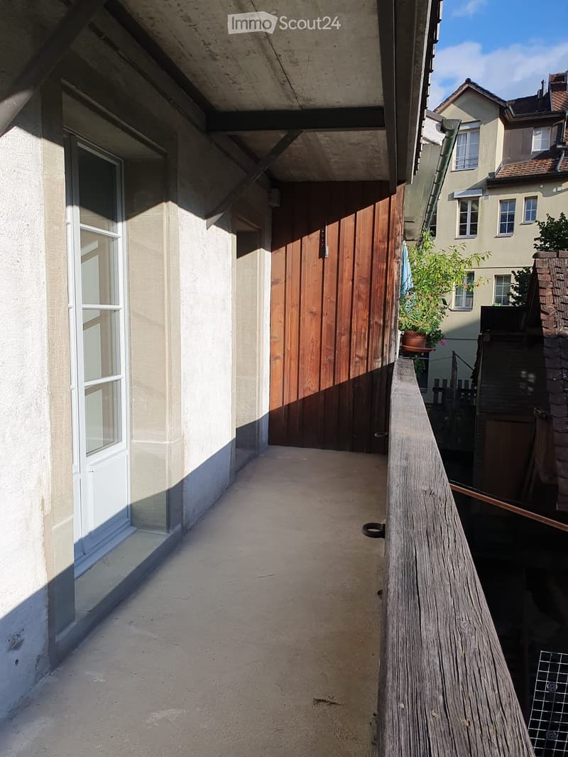 Balcony with wooden railing, concrete floor, and a wooden door leading to the building.