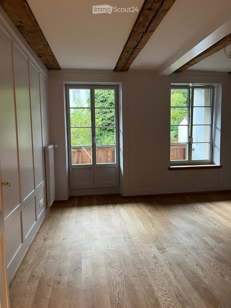 Empty attic room with wooden flooring, exposed wooden beams, large windows, and a door leading outside.
