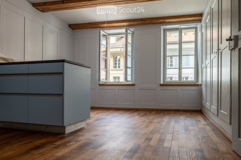 An unfurnished room with light grey walls, wooden flooring, and large windows offering a view of the street. The room features built-in cabinetry and exposed wooden beams on the ceiling.