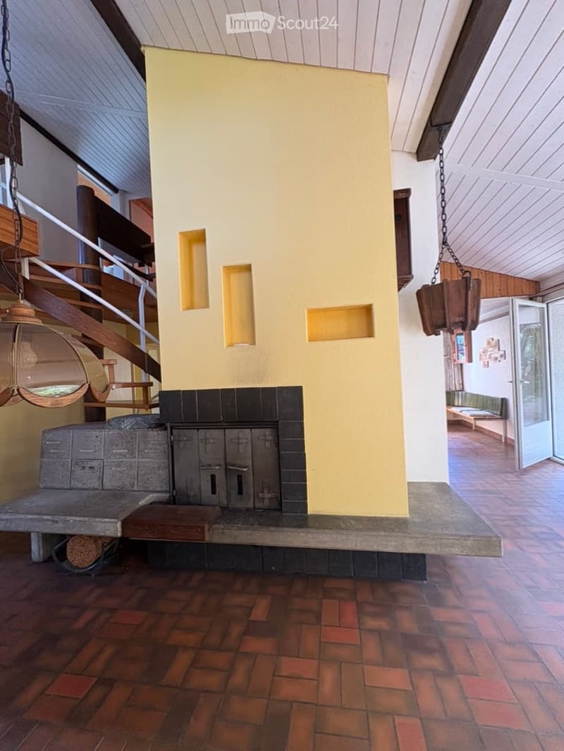 Hallway with a central fireplace, wooden ceiling, and hanging lanterns. The space features a staircase leading upwards and a bench near the fireplace.