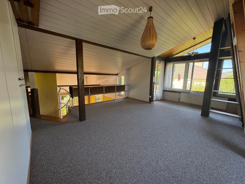 Empty room with carpeted floor, wooden ceiling beams, large windows, and a staircase leading to another level.