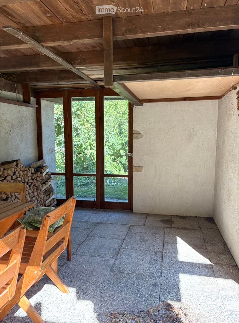 A rustic, unfurnished room with a wooden ceiling, tiled floor, and a stack of firewood near a fireplace. A wooden chair and table are positioned near the entrance.