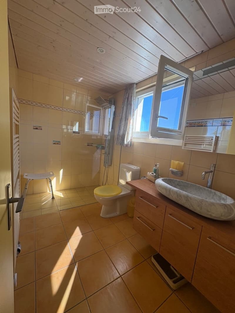 Bright bathroom with wooden slanted ceiling, tiled walls and floor, large window, toilet with yellow seat, sink with wooden cabinet, and a small stool near the shower area.