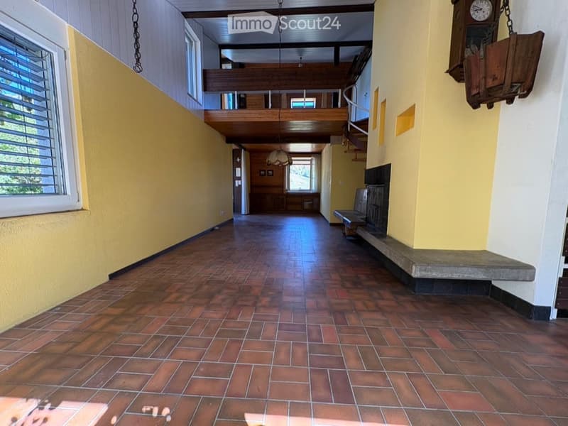 Spacious hallway with wooden flooring, yellow walls, and a staircase leading to an upper level. Features include a bench, a hanging clock, and large windows allowing natural light.