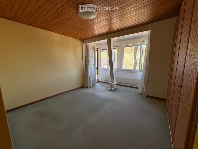 Empty room with wooden ceiling, carpeted floor, large windows with curtains, and a small white stand in the corner.