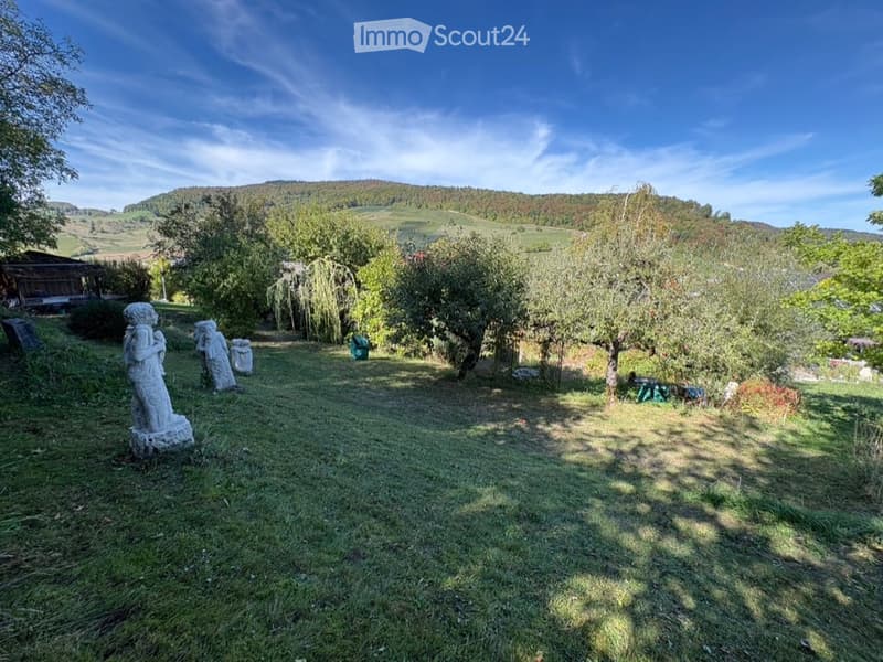 A serene garden area with grassy lawn, scattered stone statues, fruit trees, and distant vineyard hills under a clear blue sky.