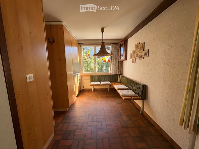 A cozy, narrow room with wooden flooring, a built-in bench seating area, and a small cabinet. The room features a hanging light fixture, a window with curtains, and several picture frames on the wall.