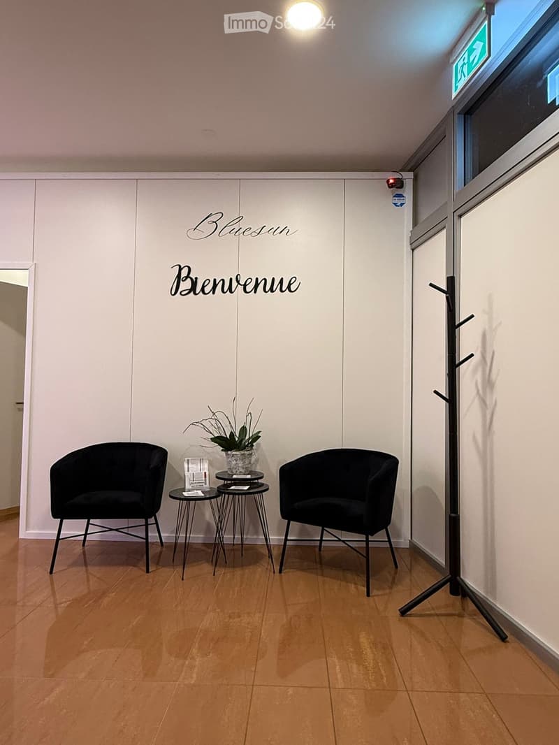 A small, modern waiting area with two black chairs, a small round table with a plant, and a coat rack. The walls are white with a welcome sign.