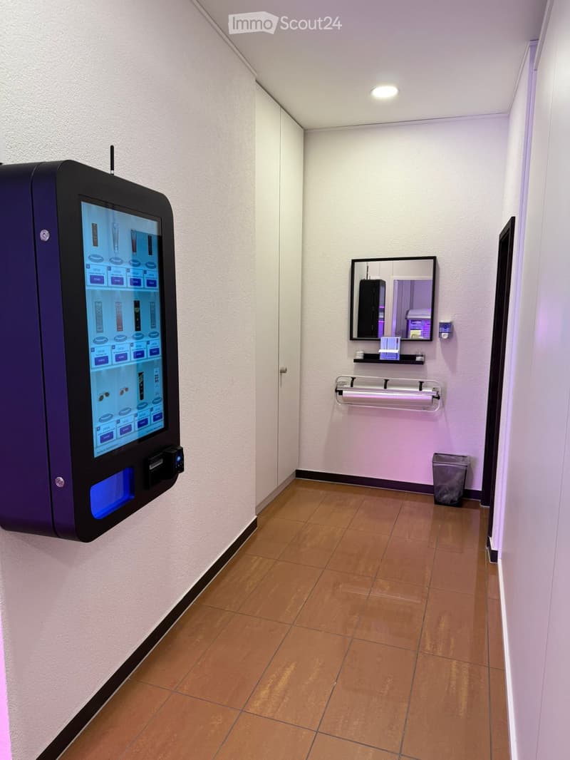 A narrow hallway with light-colored walls, a tiled floor, and a digital vending machine on one side. A small mirror, shelf, and trash bin are visible on the opposite wall.