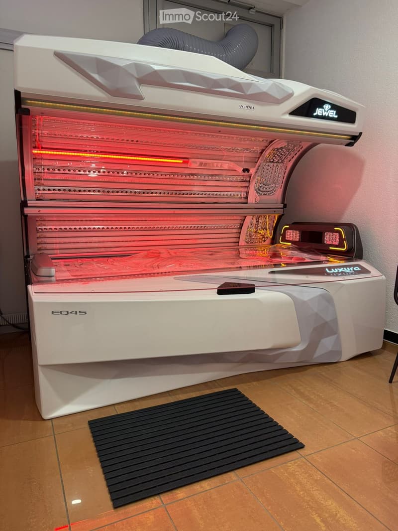Modern sunbed with control panel, placed on a wooden floor with a black mat in front.