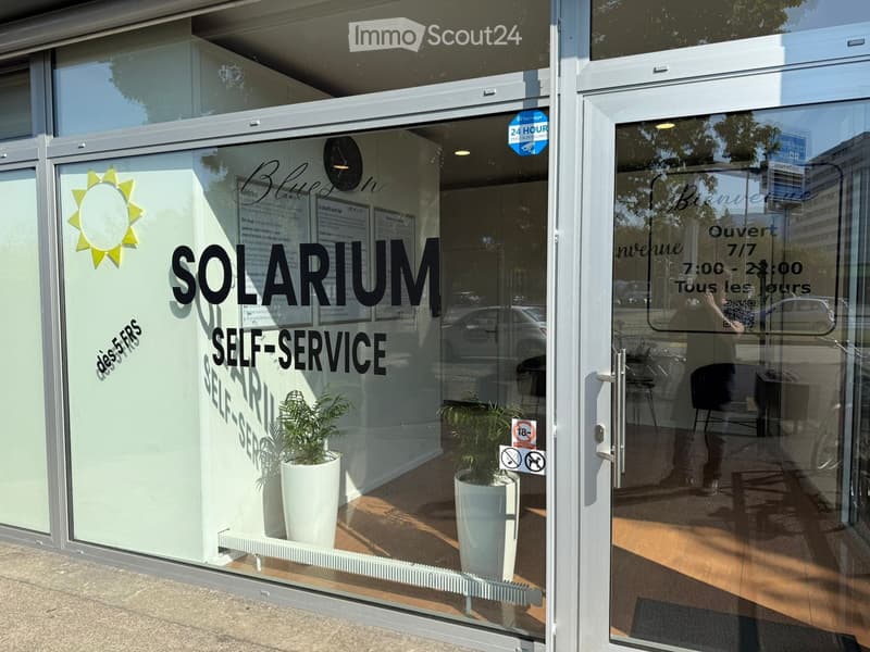 Storefront with glass windows and doors, signage reading "SOLARIUM SELF-SERVICE", potted plants inside, and a clock on the wall.