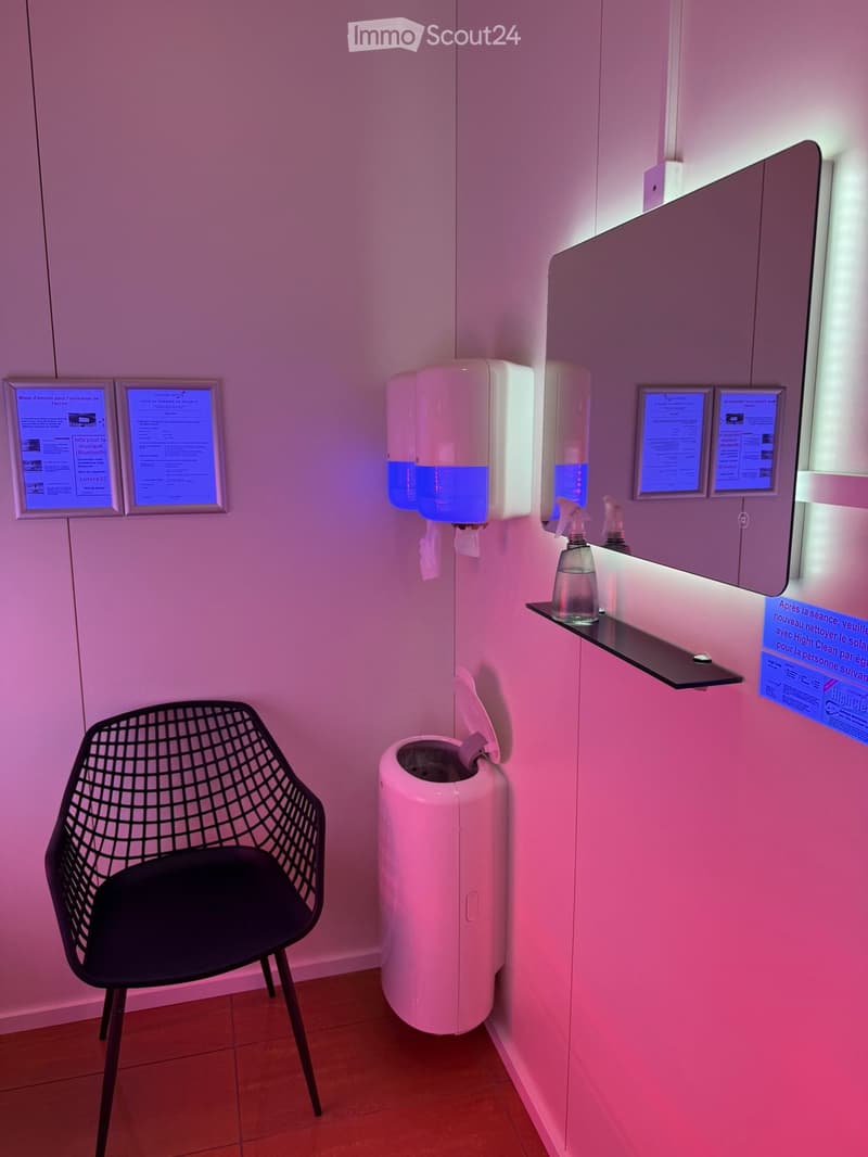 Modern bathroom with a black wireframe chair, hand sanitizer dispensers, a mirror with a built-in screen, and a trash can.