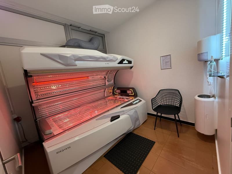 A small, well-lit room with a large sunbed, a black wire chair, a trash can, and a hand sanitizer dispenser on the wall.