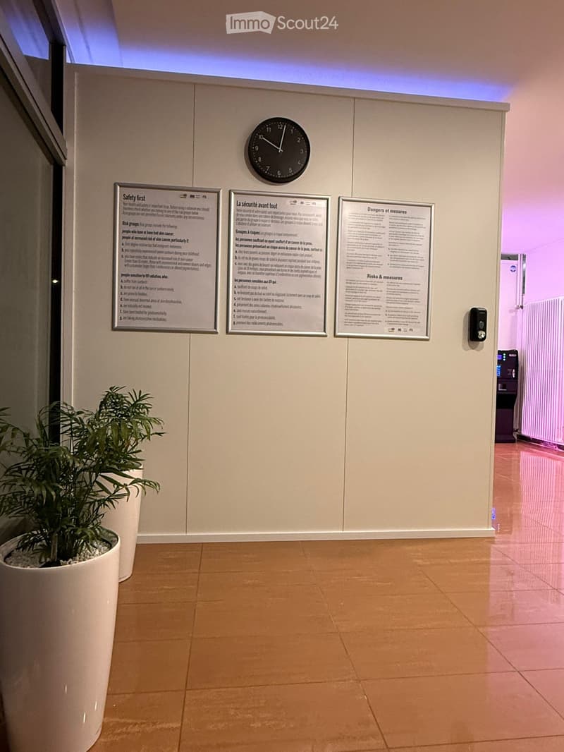 Hallway with beige walls, wooden floor, potted plant, informational posters, and a wall-mounted clock.