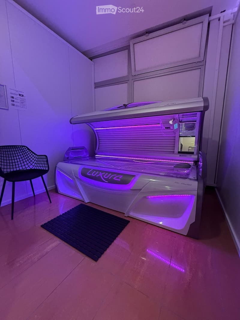 A small, well-lit room with a modern tanning bed and a single chair, featuring a sleek design and purple lighting accents.