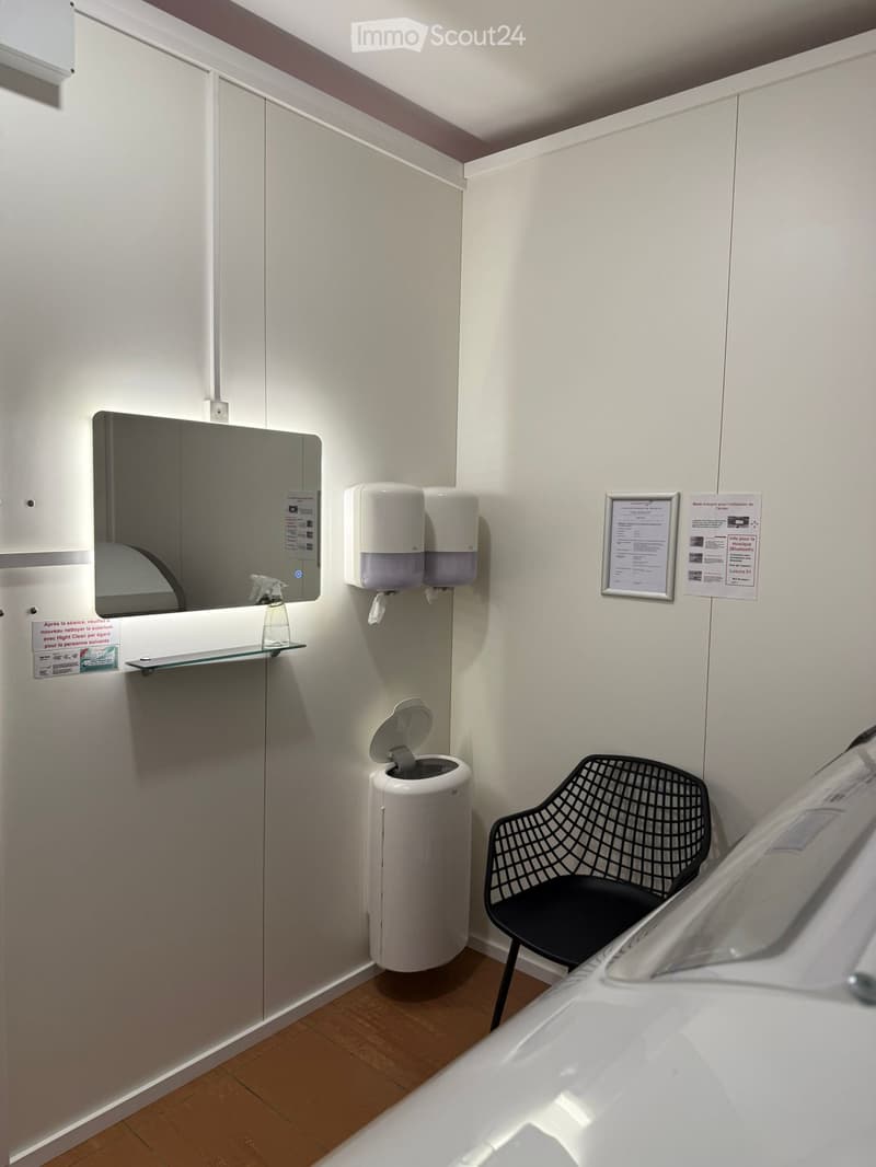 Small, modern room with a mirror, hand sanitizer dispenser, trash can, and a black wire chair.