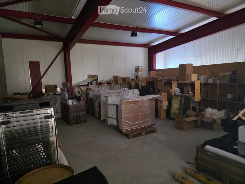 An empty warehouse or storage space with exposed metal beams, corrugated metal walls, and scattered boxes and items, including a large wrapped object, shelves, and a fan.