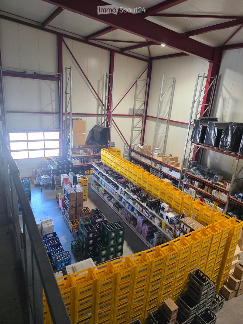 Warehouse with high ceilings, metal shelving units, and numerous yellow crates stacked on the floor. Shelves hold bottles and boxes, with a large window allowing natural light to enter.