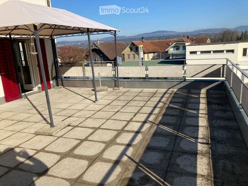 Balcony with concrete tiles, metal railing, and a large umbrella providing shade. The background shows a view of a town with houses and mountains.