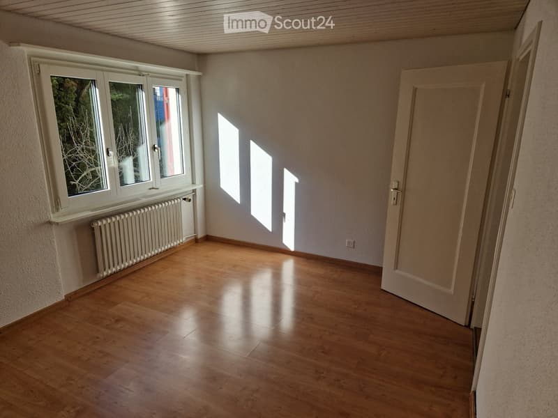 Empty room with wooden flooring, white walls, and a window with a radiator below it. Sunlight streams in from the window, casting shadows on the floor.