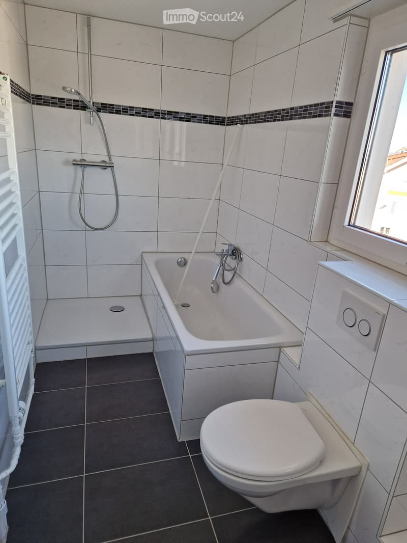 Modern bathroom with a bathtub, toilet, showerhead, and a window providing natural light. The walls are tiled with a decorative border, and the floor is dark tiled.