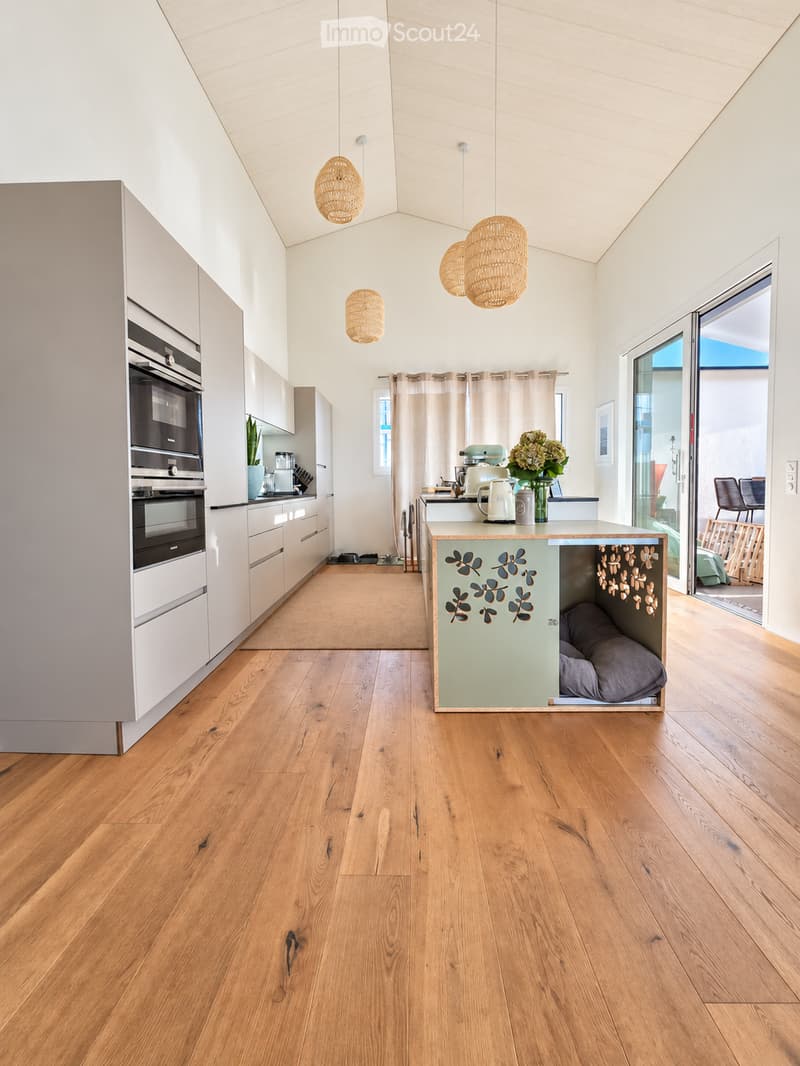 Modern kitchen with light-colored cabinets, wooden flooring, pendant lights, and a kitchen island with a built-in pet bed.