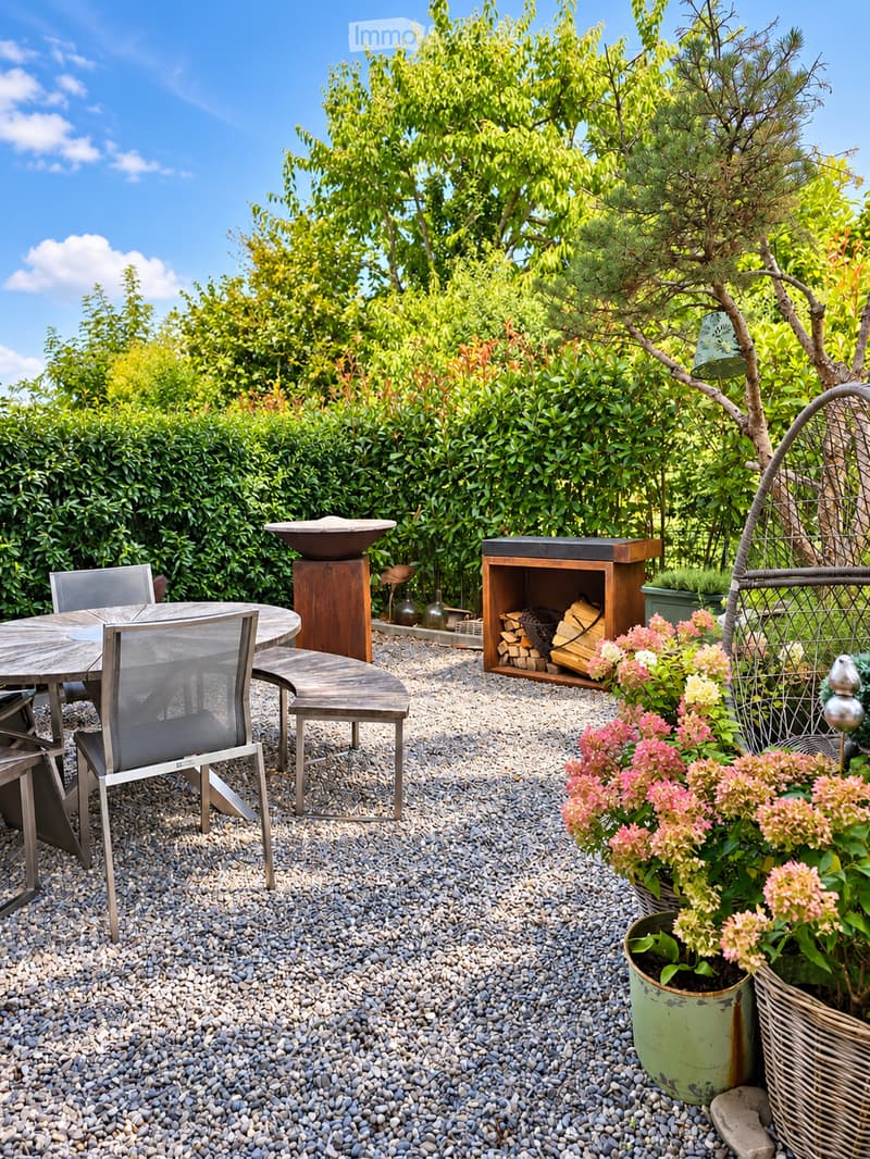 A well-maintained garden area with a round wooden table and chairs, a wooden bench, a decorative wooden storage box, a stone bowl, and various potted plants and flowers.