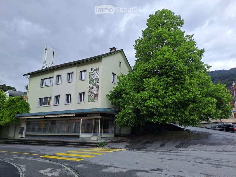 Two-story building with light yellow facade, multiple windows, and a mural on one side. The building has a small awning over the entrance and is surrounded by trees and a road with a crosswalk.
