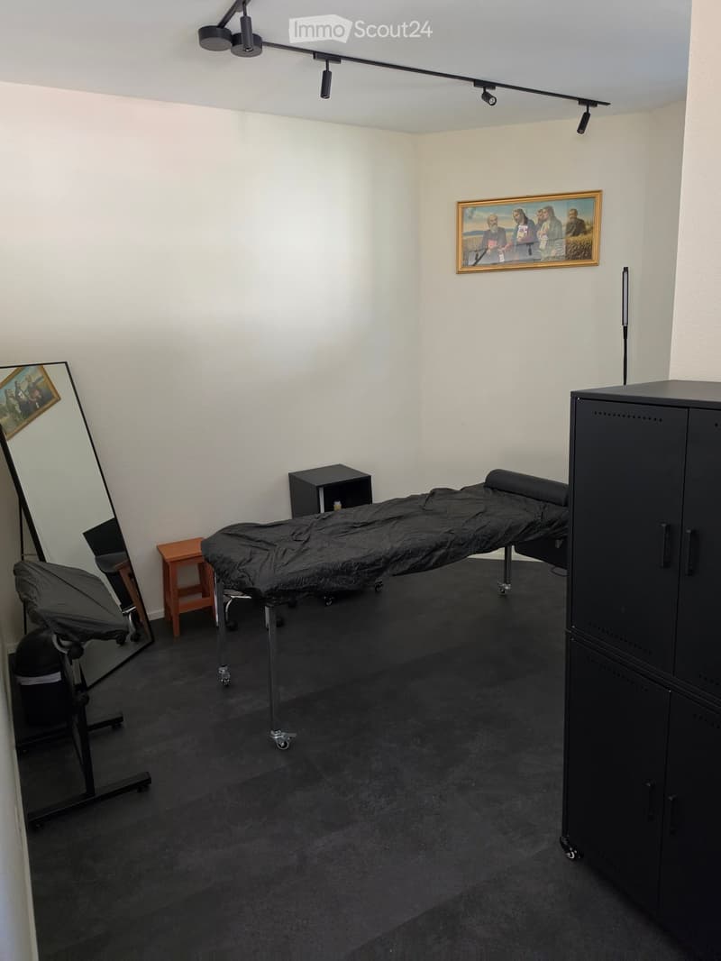 A well-lit room with a massage table, a black cabinet, a small wooden stool, a black chair, and a mirror with a picture frame on the wall.