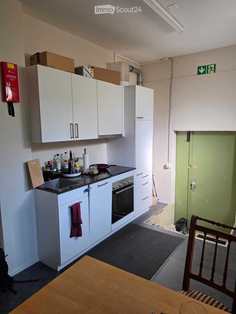 A small kitchen with white cabinets, a sink, an oven, and a wooden dining table with a chair. Various kitchen items are on the counter, and there is a fire extinguisher on the wall.