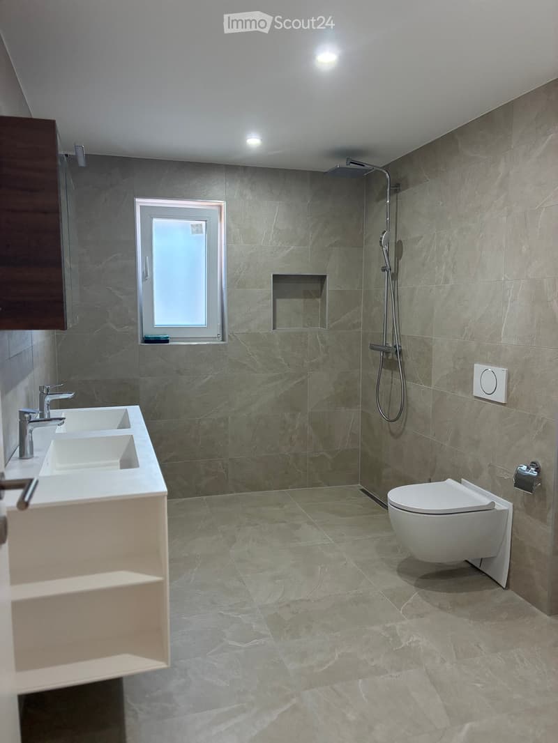 Modern bathroom with two sinks, a toilet, a shower, and a window with frosted glass. The walls and floor are covered in grey marble tiles.
