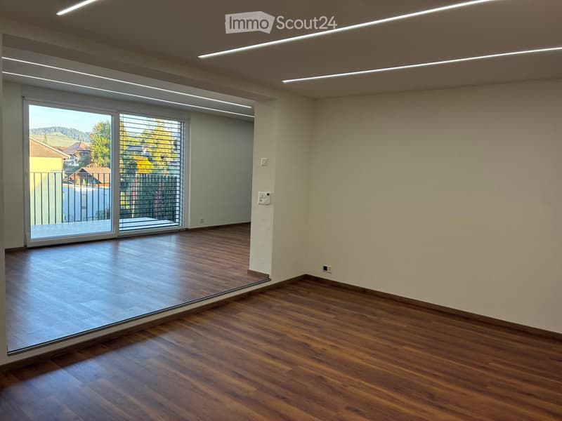 Empty room with wooden flooring, large sliding glass door leading to a balcony, and modern lighting fixtures on the ceiling.