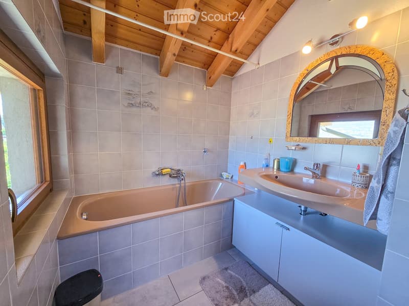 Bathroom with a bathtub, double sink, mirror, and toilet. Features wooden beams on the ceiling and a large window letting in natural light.