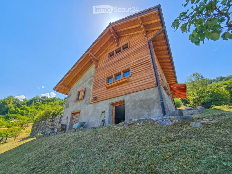 A two-story wooden house with a stone base, situated on a grassy slope with surrounding trees and a clear blue sky.