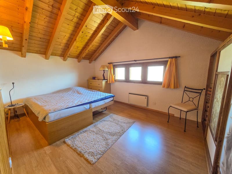 Cozy attic bedroom with wooden slanted ceilings, a bed, a small table with a lamp, a chair, and a window with curtains.