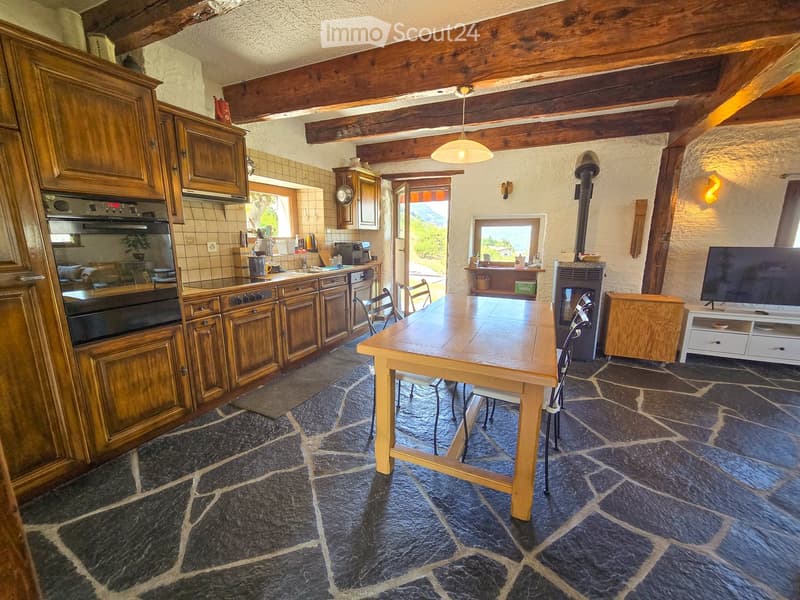 A rustic kitchen and dining area with wooden cabinets, a stone-tiled floor, a wooden dining table with chairs, and a television on a stand.