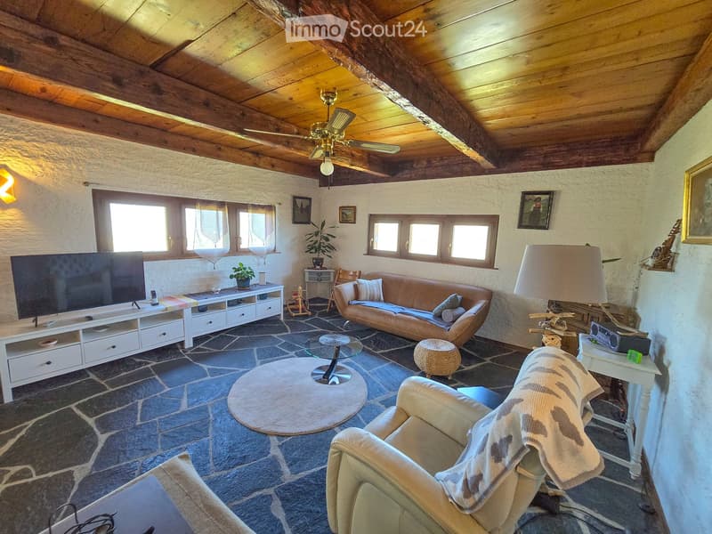Living room with exposed wooden beams, stone flooring, a brown leather sofa, a white armchair, a coffee table, a TV stand, and a table lamp.