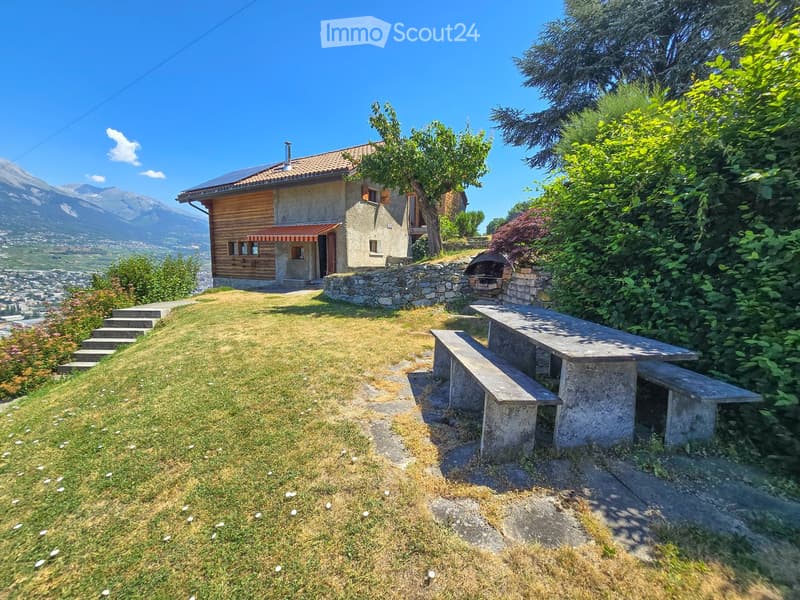 A grassy outdoor area with a stone picnic table and benches, surrounded by trees and shrubs, with a wooden house and stone wall in the background and a scenic mountain view.