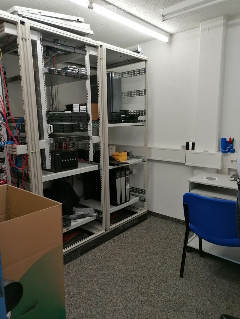 A server room with a large open server rack, a blue chair, a white desk, and various cables and equipment.