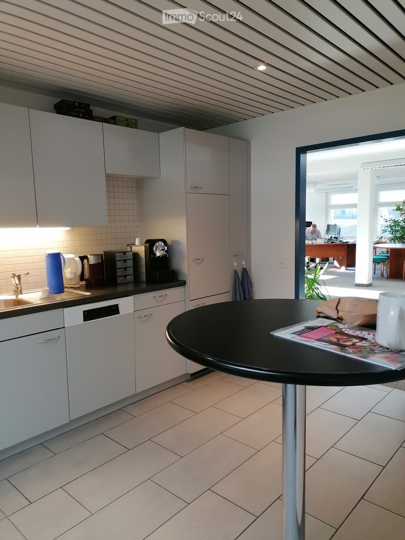 Modern kitchen with white cabinets, tiled backsplash, and a black countertop. A small round table with a mug and magazine is visible, and a coffee maker and other appliances are on the counter.