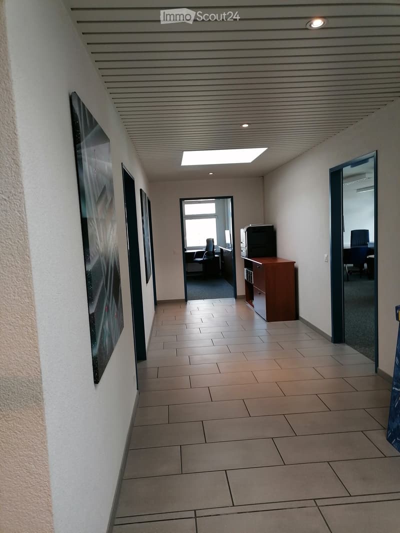 A clean, modern hallway with tiled flooring, white walls, and recessed lighting. There are framed artworks on the left wall and a small office area visible at the end of the corridor.