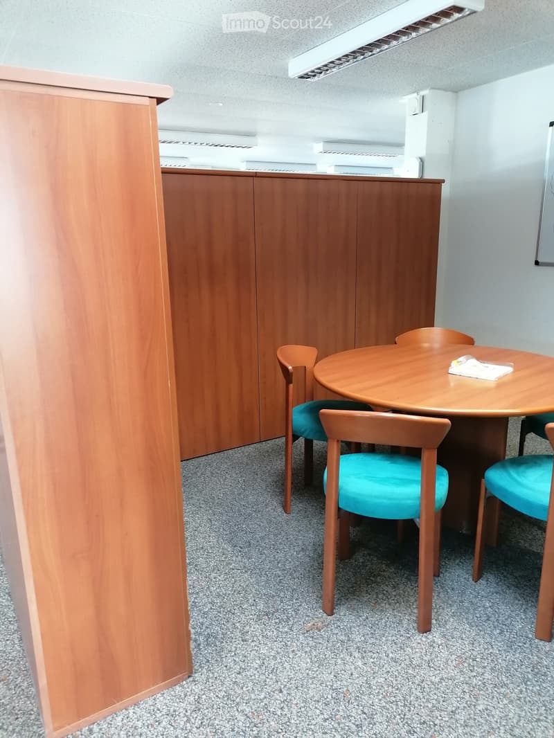 A small, furnished room with a round wooden table and four matching chairs with teal cushions, set against a backdrop of wooden cabinetry and a grey carpeted floor.
