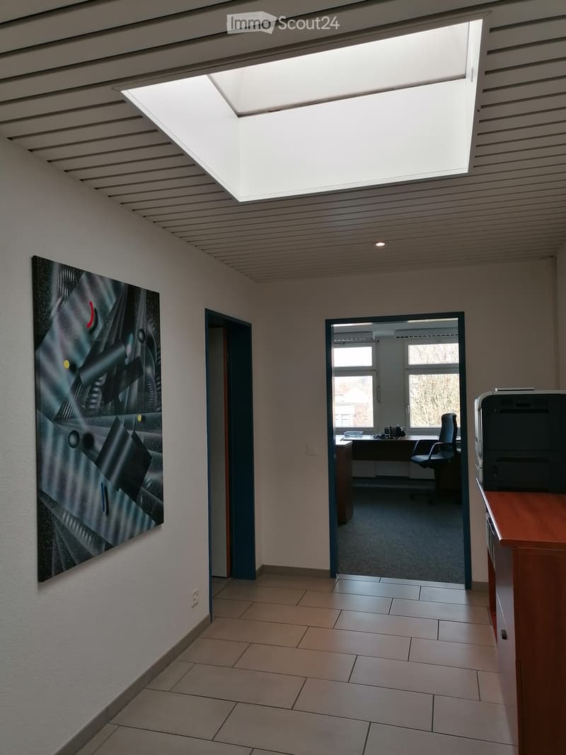 Modern hallway with tiled floor, white walls, large abstract artwork, and a partially visible office area with a desk and chair.