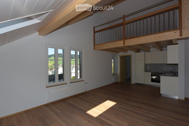 Bright, open-plan living and dining area with wooden flooring, large windows offering a scenic view, and a modern kitchen area in the background.