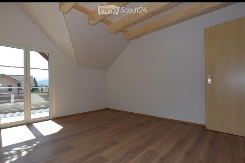 Empty room with wooden flooring, white walls, and a slanted wooden ceiling. Features a large window with a view of the outside and a wooden door.